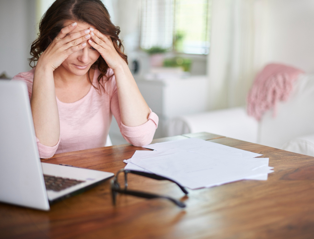 Stressed women with hands on her head at wooden desk with papers laptop and glasses Stressed women with hands on her head at wooden desk with papers laptop and glasses