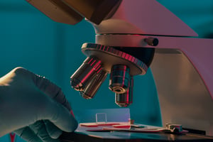 scientist wearing a white glove testing a sample under a microscope with a teal background
