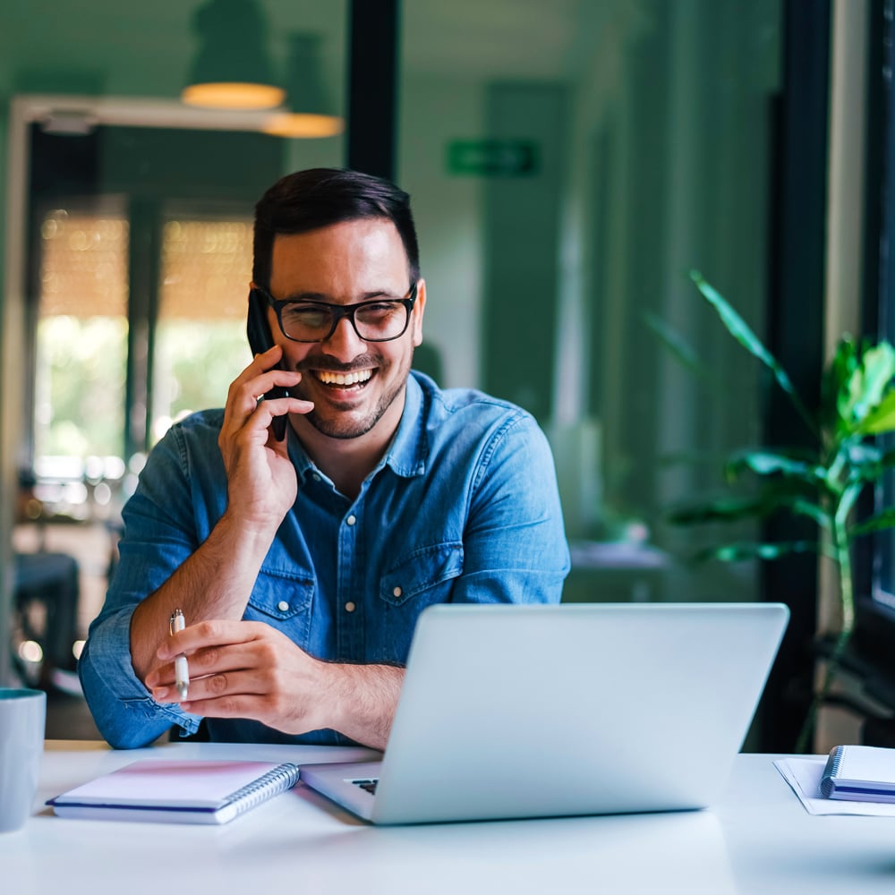 Portrait-of-young-smiling-cheerful-clinical-study-participant-in-casual-office-receiving-phone-call-from-Atlantia-recruitment-officer-after-signing-up-for-a-study-on-laptop