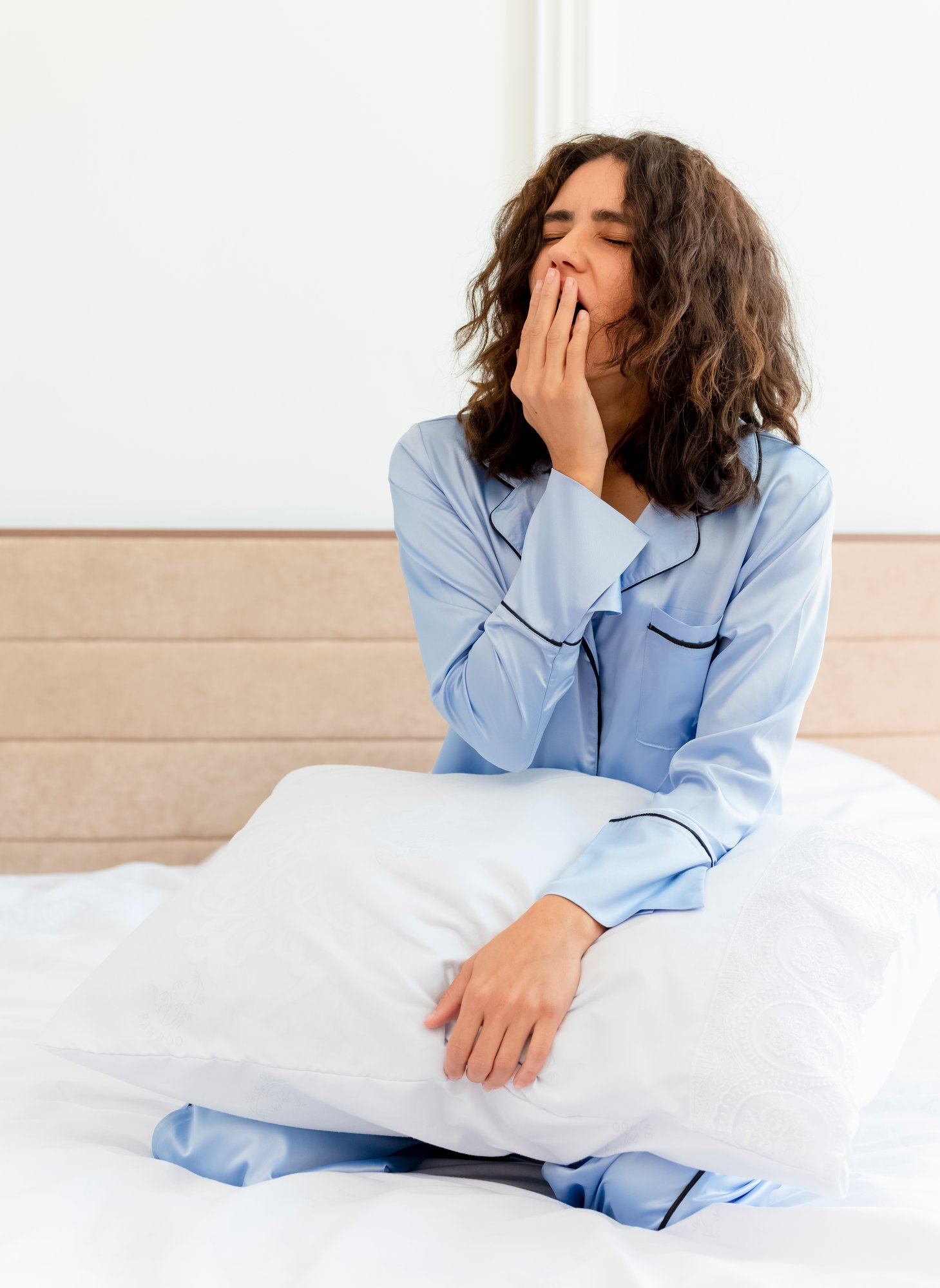 young-beautiful-woman-blue-pajamas-sitting-bed-with-pillow-waking-up-yawning-bedroom-interior-light-background